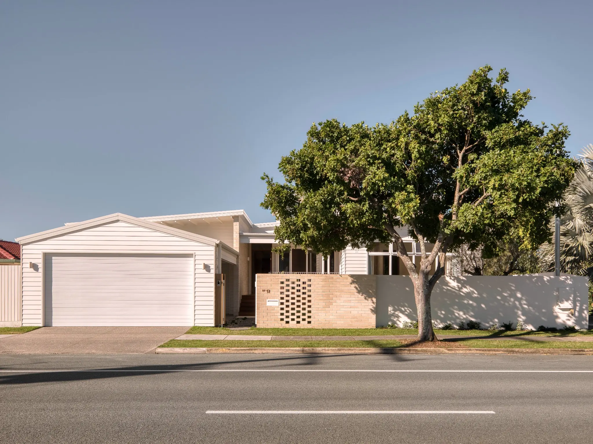 Single-story modern house with a white garage door, beige brick wall, and a large green tree in front, under a clear blue sky.