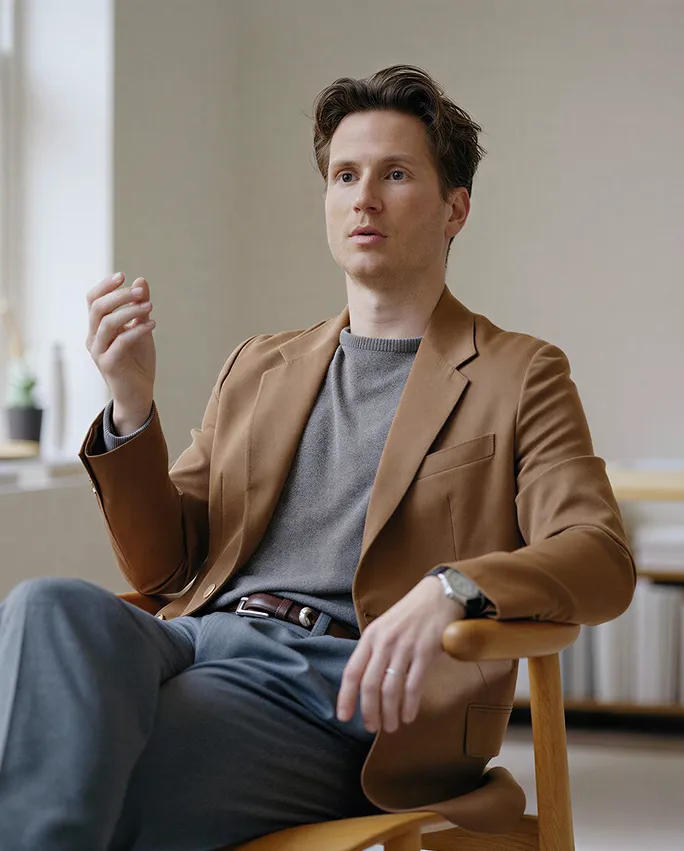 Man in brown blazer and gray sweater gesturing while sitting on wooden chair in a modern office.