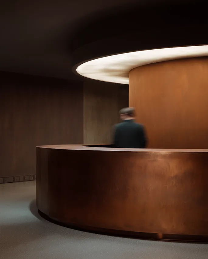 Blurred figure stands behind a curved brown reception desk under soft circular ceiling light.