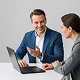 Two business professionals in a discussion, one pointing at a laptop screen, both smiling.