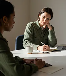 Two women having a thoughtful discussion at a table with notebooks and a coffee cup.