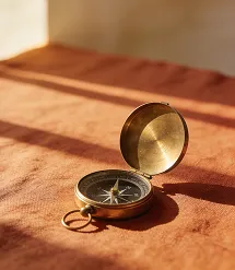 Open brass compass resting on textured brown fabric under warm sunlight.