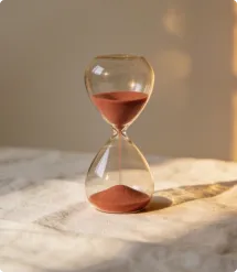 Hourglass with red sand partially filled on a fabric surface, casting a shadow.