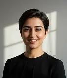 Smiling woman with short dark hair wearing a black top, with sunlight and shadow on the wall behind her.
