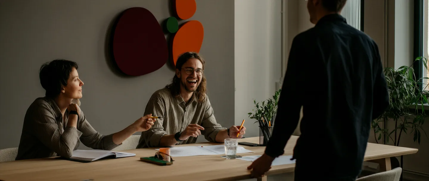 Two seated colleagues smiling and talking to a standing man during a meeting at a table with papers.