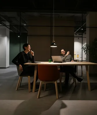 Two people sitting at a wooden table in a modern office, smiling and engaged in conversation, with a laptop and a green cup on the table, illuminated by a single overhead pendant light.