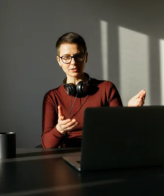 Person wearing glasses and headphones around the neck, gesturing with hands while looking at a laptop on a table in a dimly lit room with shadows on the wall.