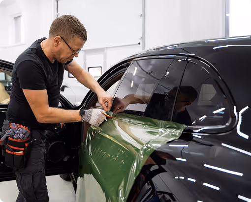 Man wearing glasses and gloves applying green vinyl wrap to the door of a black car in a workshop.