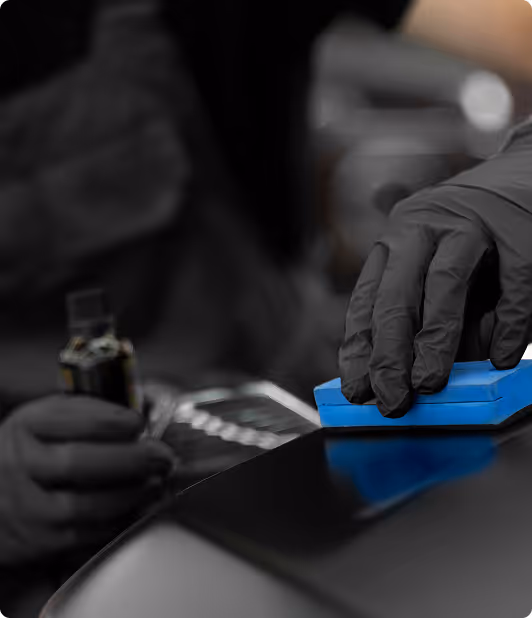 Person wearing black gloves applying a product with a blue sponge on a shiny black surface.