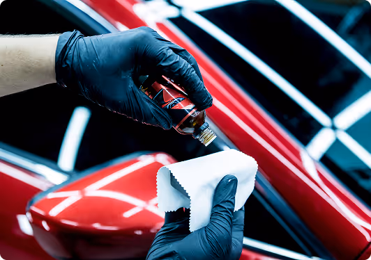 Person wearing black gloves pouring liquid from a small bottle onto a white cloth near a red car's side mirror.