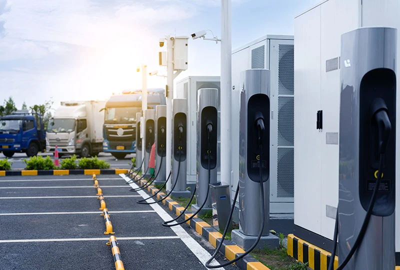 Row of modern electric vehicle charging stations with cables hanging, situated on a paved lot with empty parking spaces and trucks in the background under a partly cloudy sky with sunlight.