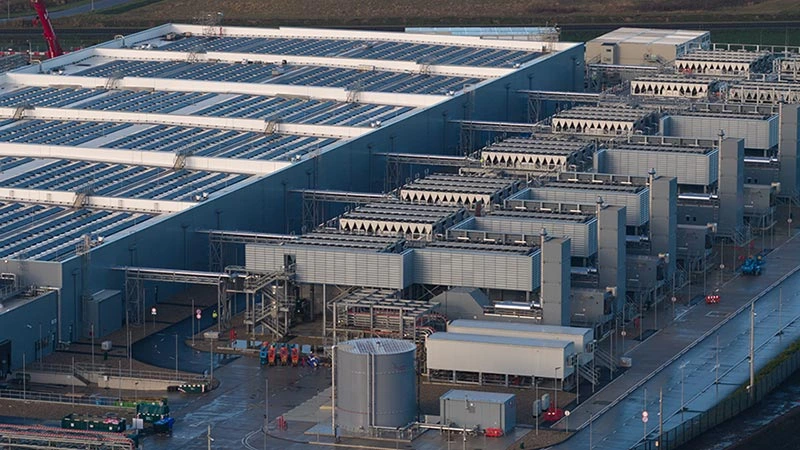 Large data center facility with multiple cooling units and HVAC systems lined up beside a warehouse with a solar panel-covered roof.