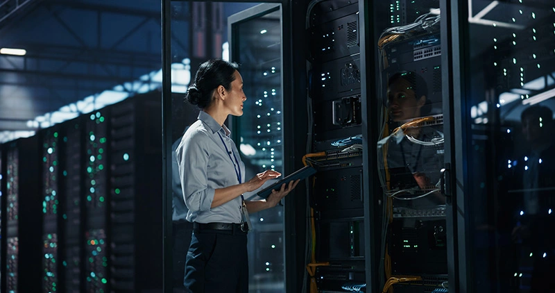 Woman in business attire holds a tablet while inspecting a server rack in a dimly lit data center with rows of servers.