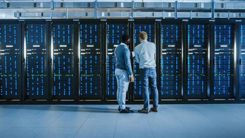 Two men standing and looking at multiple server racks filled with computers and blue LED lights in a data center.