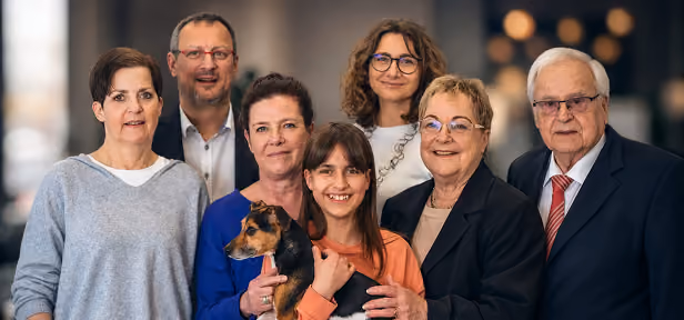 Eine fröhliche Gruppe von sechs Personen verschiedenen Alters, darunter eine junge Frau mit braunen Haaren, die einen kleinen Hund in den Armen hält; alle blicken lächelnd in die Kamera, vor einem unscharfen, dunklen Hintergrund.