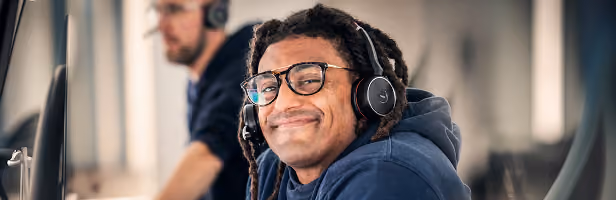 Junger Mann mit Dreadlocks, der Kopfhörer und Brille trägt, lächelt in einer modernen, gut beleuchteten Büroumgebung mit einem verschwommenen Kollegen im Hintergrund.