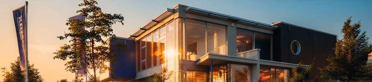 Modernes Haus mit großen Glasfenstern, das von Bäumen umgeben ist, bei Sonnenuntergang mit warmem Licht und blauem Himmel.