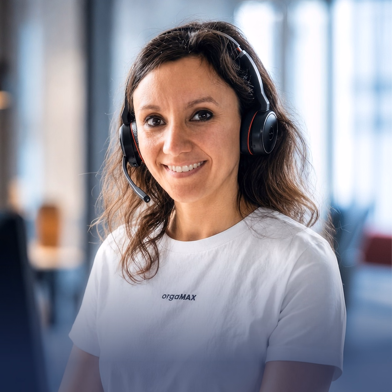 Lächelnde Frau mit dunklen Haaren trägt ein weißes T-Shirt mit dem Aufdruck "orgaMAX" und ein Headset mit Mikrofon in einem modernen Büro.