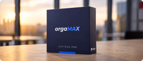 Dunkelblaue Produktbox mit der Aufschrift orgaMAX und Auftrag Pro steht auf einem Holztisch vor einem Fenster mit Sonnenuntergang.