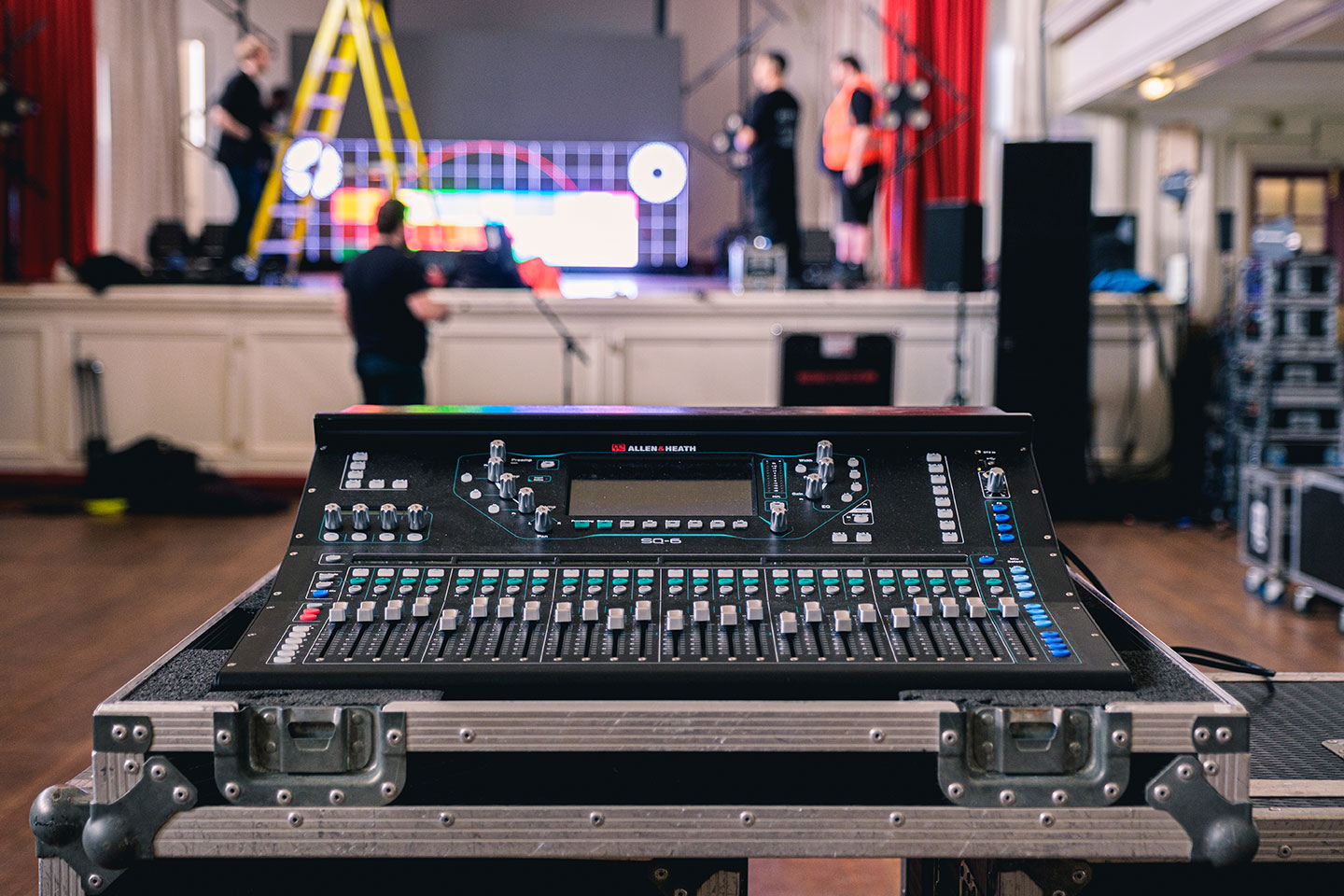 Audio mixing console in the foreground with a stage and crew blurred in background.