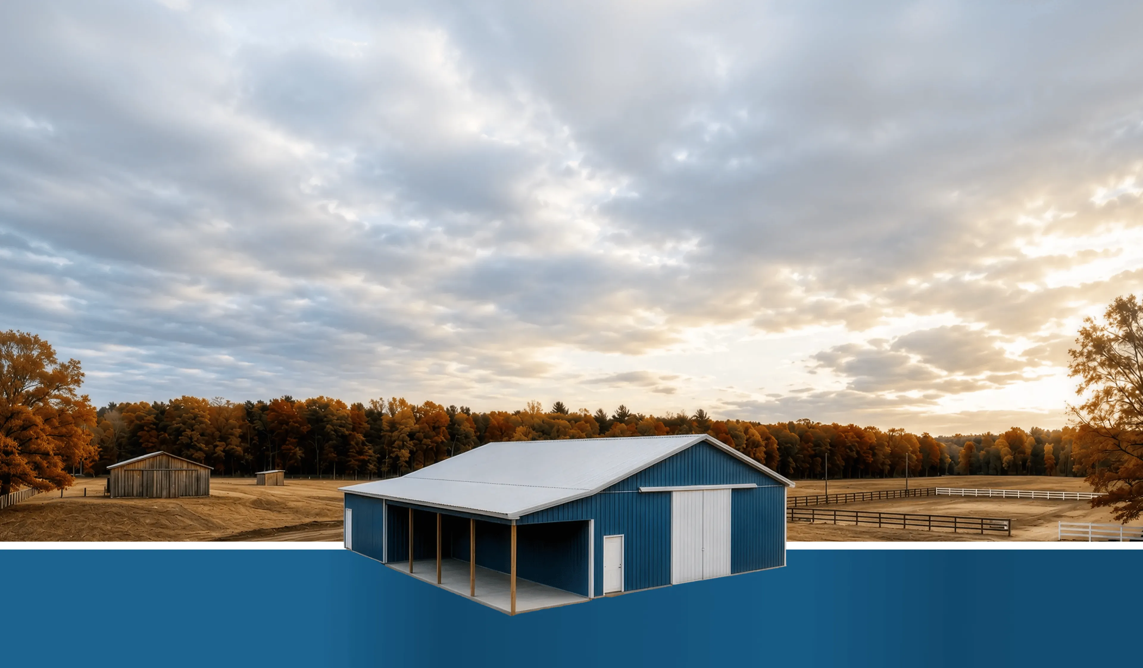 A blue barn stands in front of autumn trees and fenced fields under a cloudy sky.