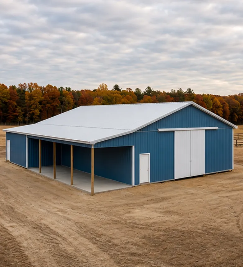 A blue and white metal barn with multiple open bays stands on bare dirt, surrounded by trees with autumn foliage under a cloudy sky.