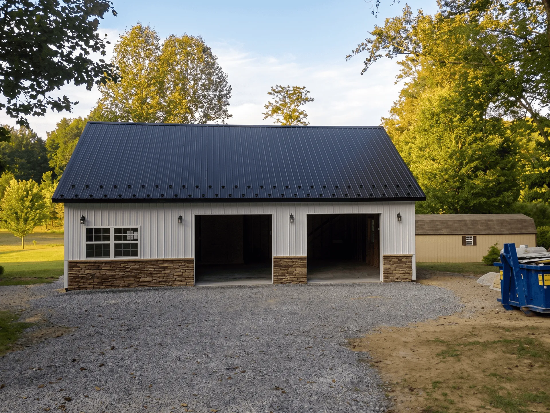 A metal garage with two open bays and one closed, stone accents, and a gravel driveway, set in a grassy yard with trees and a shed in the background.