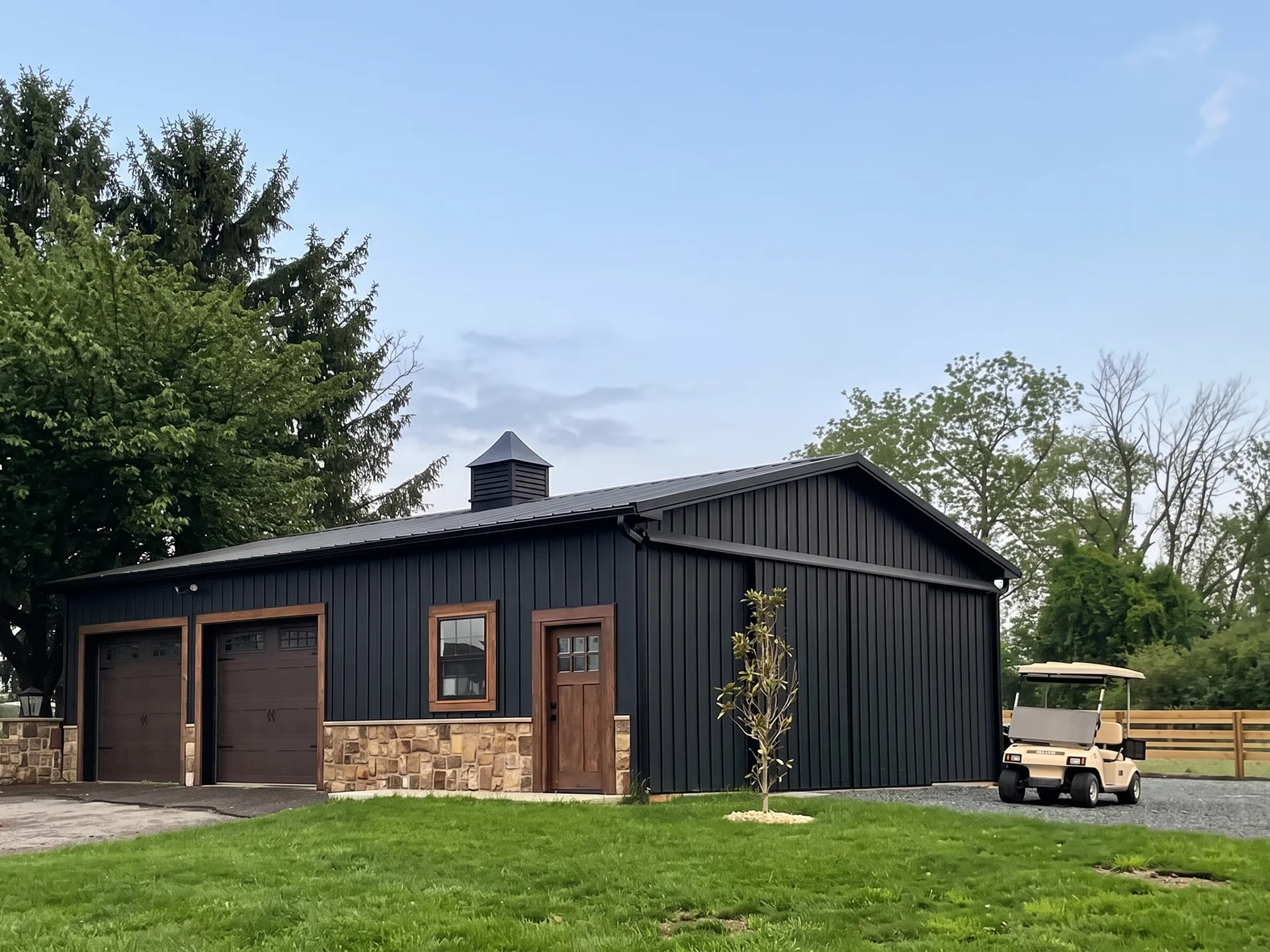 A black metal garage with stone accents, two large doors, a single door, a small tree in front, and a golf cart parked on gravel beside it.