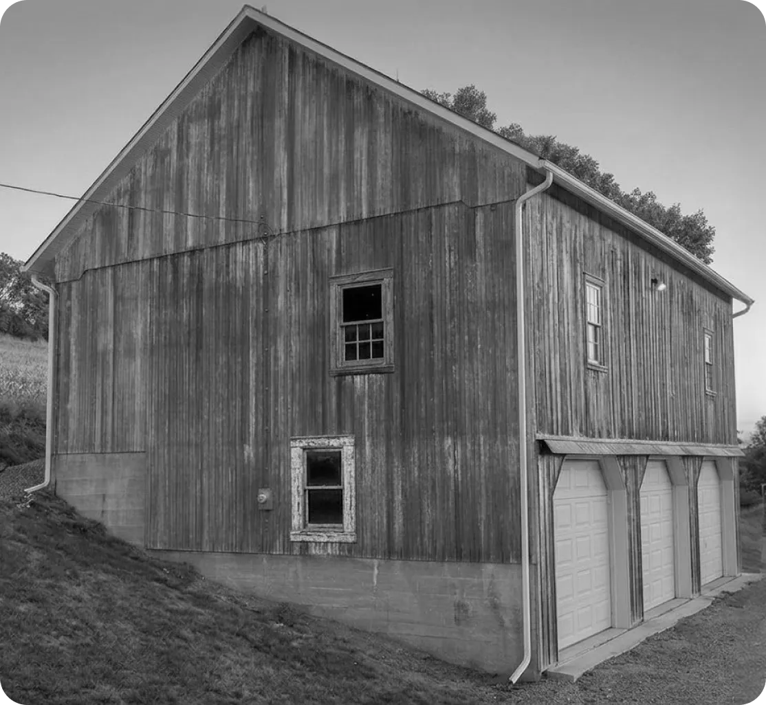 Before image of a a large wooden barn with weathered siding, two upper windows, and three white garage doors at the base.