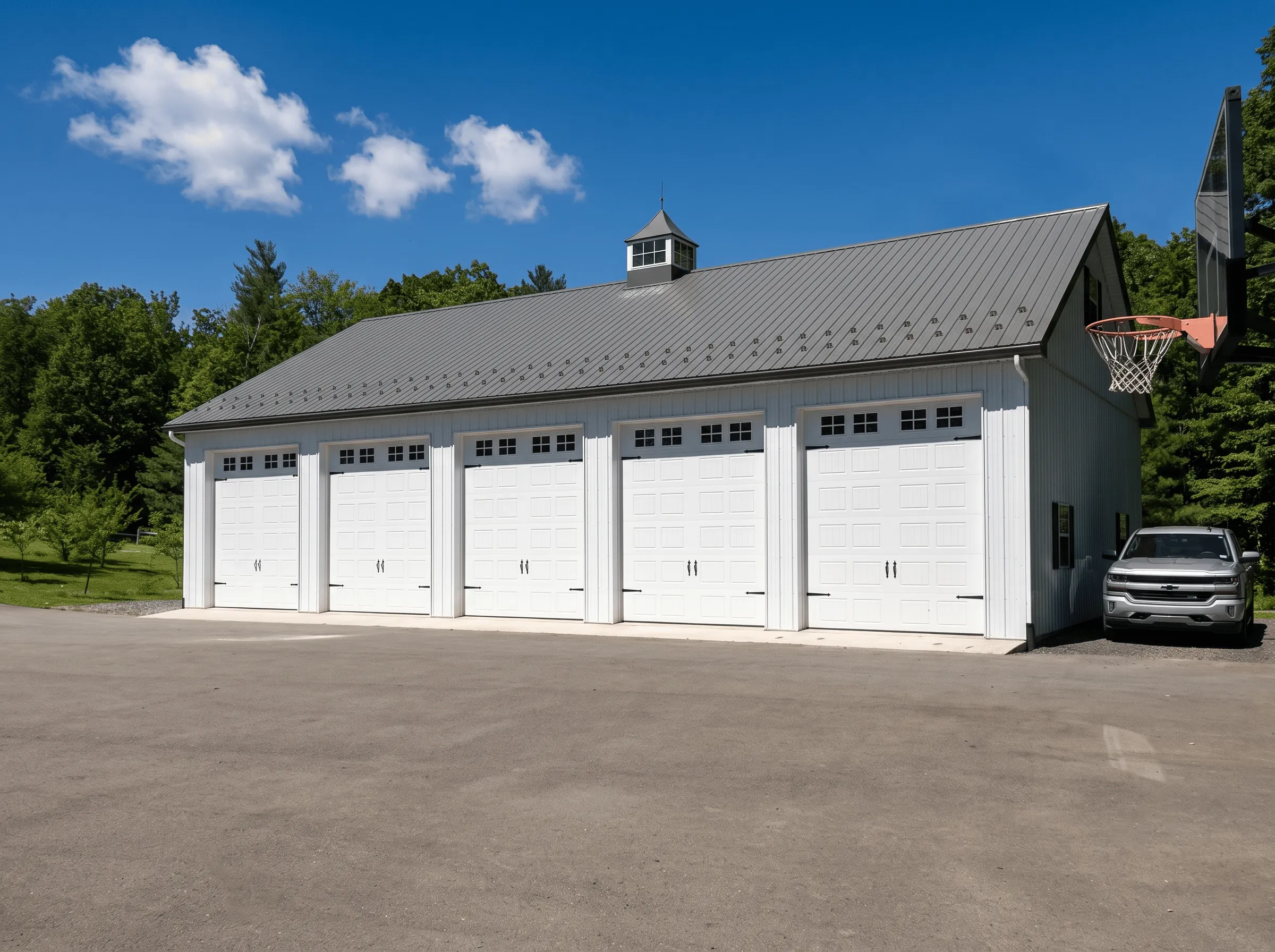 A large white garage with five closed doors, a gray metal roof, a jeep parked to the right, and a basketball hoop near the driveway.