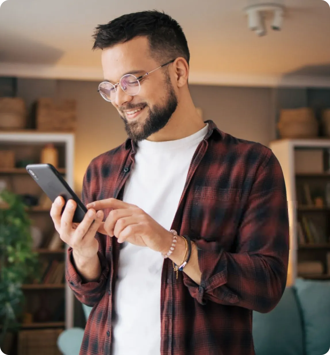 Smiling man with glasses and beard using smartphone in cozy, modern living room.
