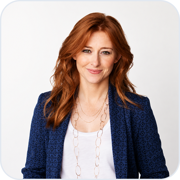 Smiling woman with long red hair wearing a blue patterned blazer over a white top and a long gold chain necklace against a white background.