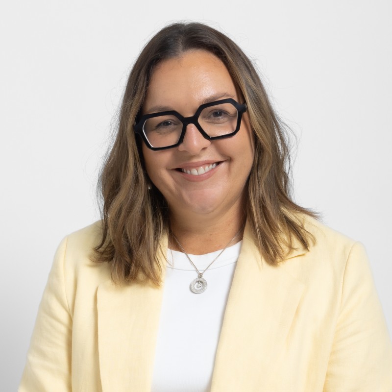 Smiling woman with shoulder-length brown hair and black geometric glasses wearing a yellow blazer, white top, and a silver pendant necklace on a plain white background.