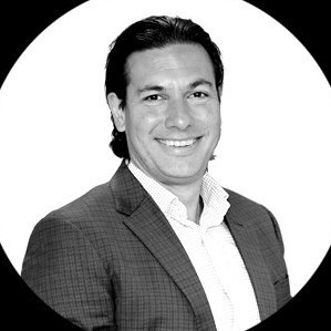 Smiling man with short dark hair wearing a checkered blazer and a white collared shirt against a white circular background.