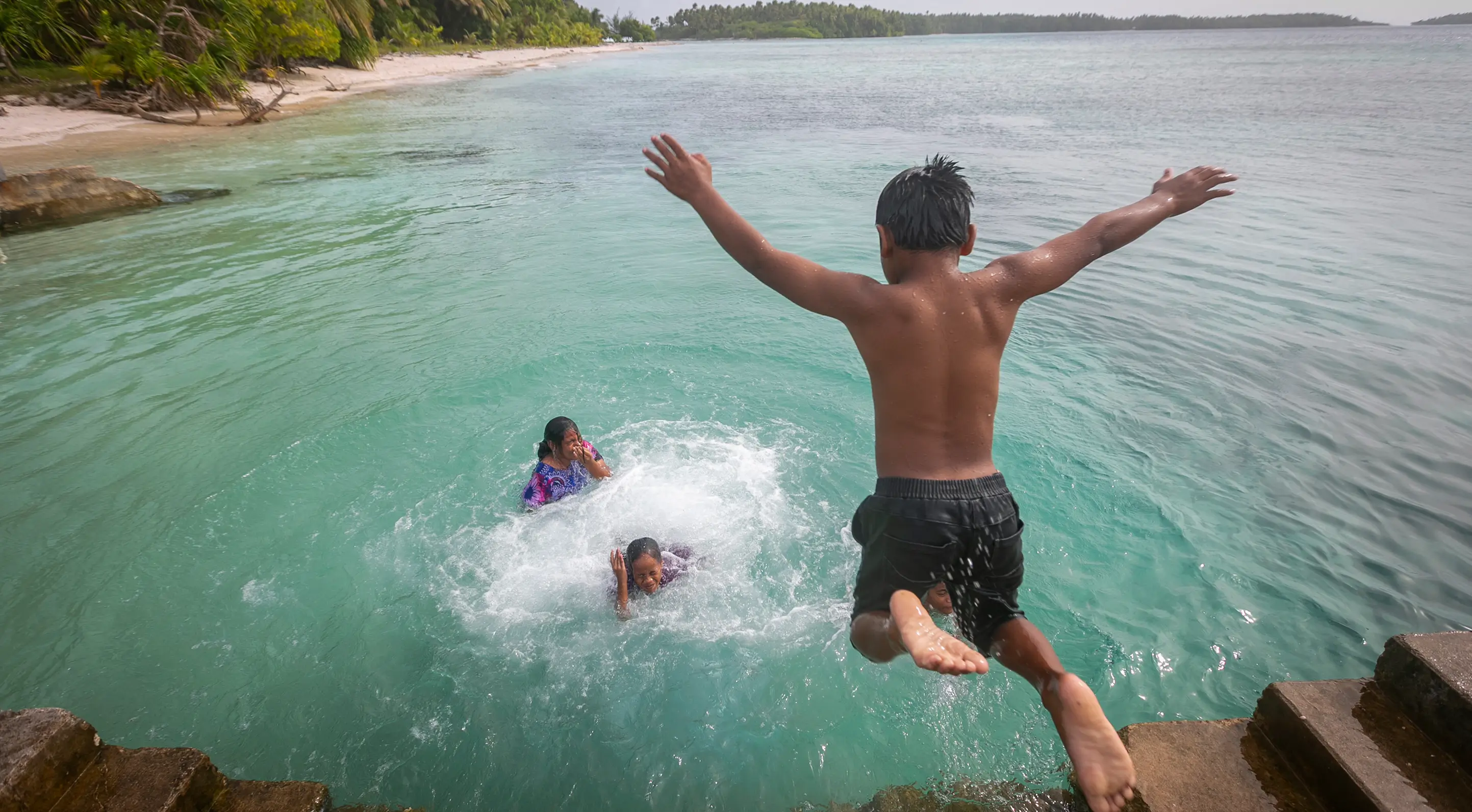 Boy jumping into clear blue water near shore where two children swim and splash.