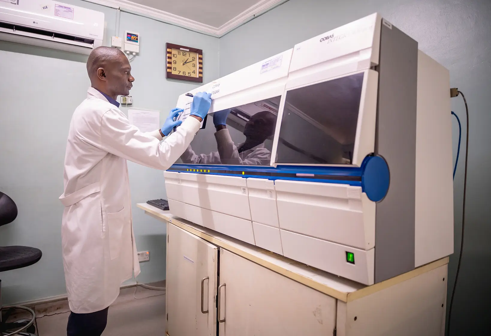 Laboratory technician in a white coat operating a large clinical analyzer machine in a lab.
