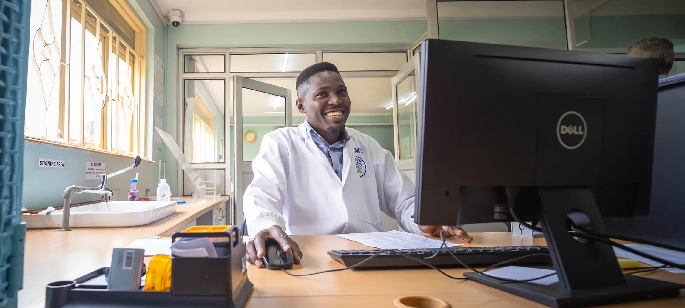 Smiling man in a lab coat working at a desk with a computer in a bright office.