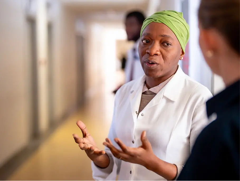 Medical professional in white coat and green headwrap talking to a colleague in a hospital hallway.