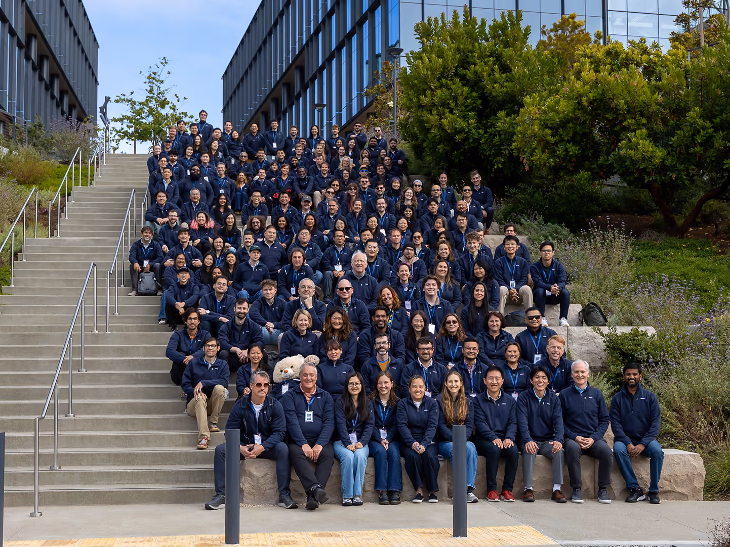 Large group of people wearing matching navy blue jackets sitting on outdoor stairs and stone steps, posing for a group photo in front of modern glass buildings and green trees.