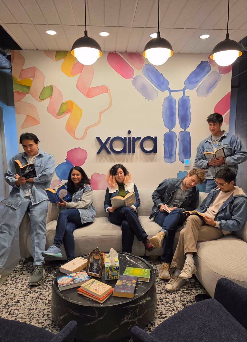 Six young adults sitting and standing around a curved beige couch, reading books, in a room with a colorful abstract wall mural and the word 'xaira' displayed on the wall behind them, illuminated by three hanging lights.