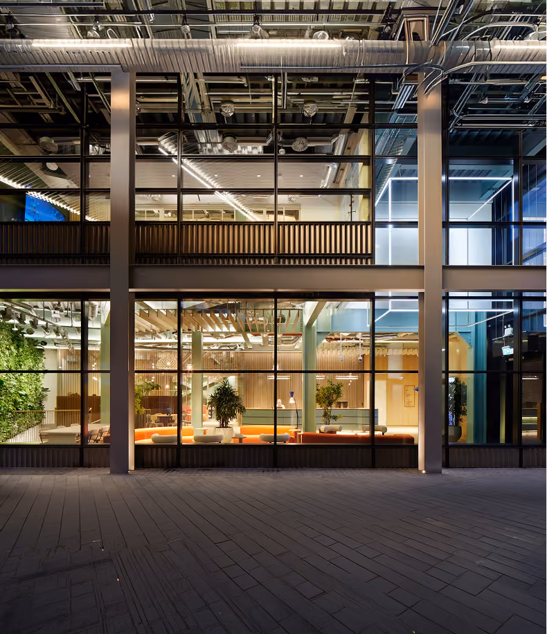 Modern building facade with large glass windows revealing a brightly lit interior featuring orange benches, potted plants, vertical wooden slats, and exposed ceiling pipes.