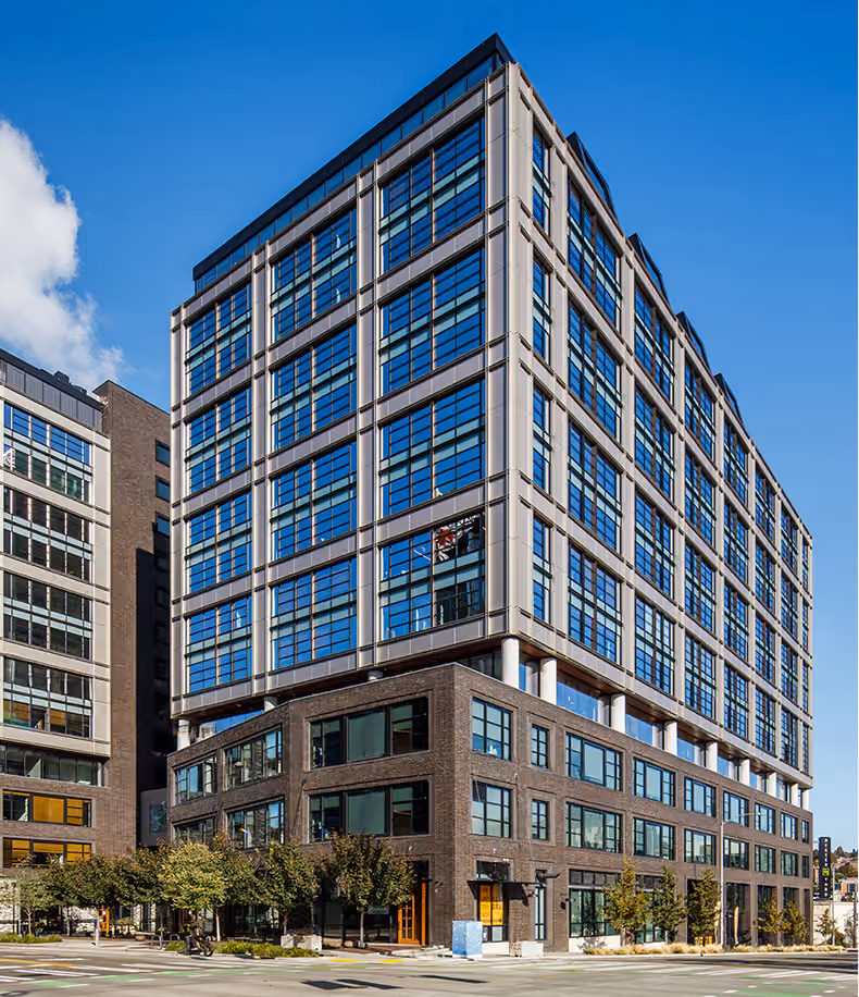 Modern multi-story office building with a lower dark brick section and a taller upper section with large blue-tinted glass windows, set against a clear blue sky.