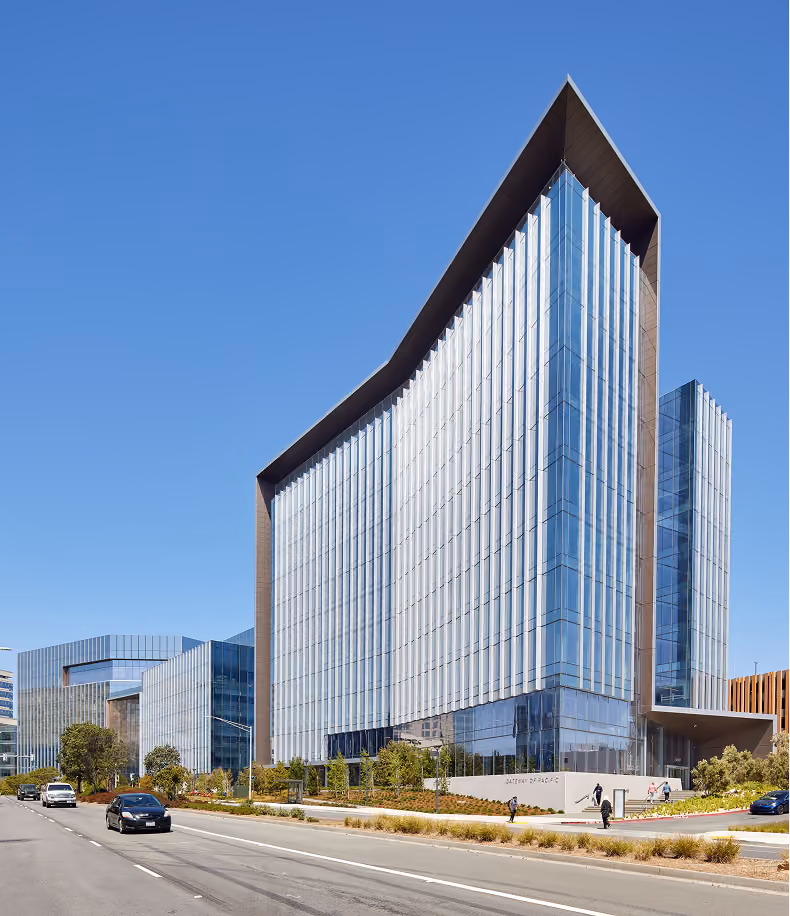 Modern glass office buildings along a multi-lane road under a clear blue sky, with cars and pedestrians on the sidewalk.