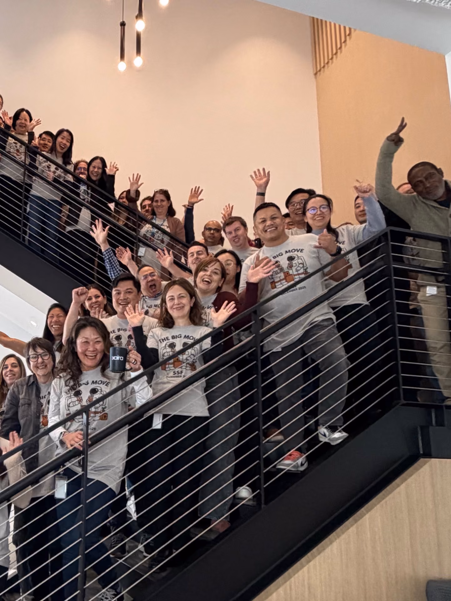 Group of smiling people standing on a staircase waving at the camera, some wearing matching 'The Big Move SSF' t-shirts.