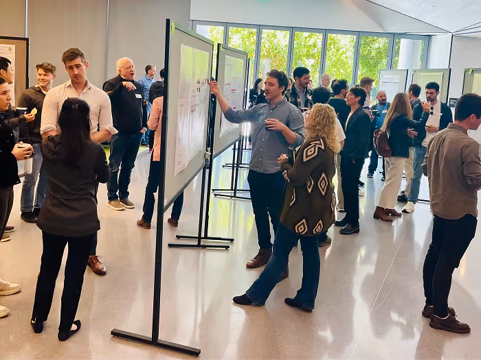 Group of people in a bright, modern indoor space attending a poster session, viewing and discussing research posters displayed on freestanding boards.