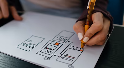 Hand holding a pen drawing a wireframe layout on paper with various boxes, lines, and colored highlights, representing website or app design planning.
