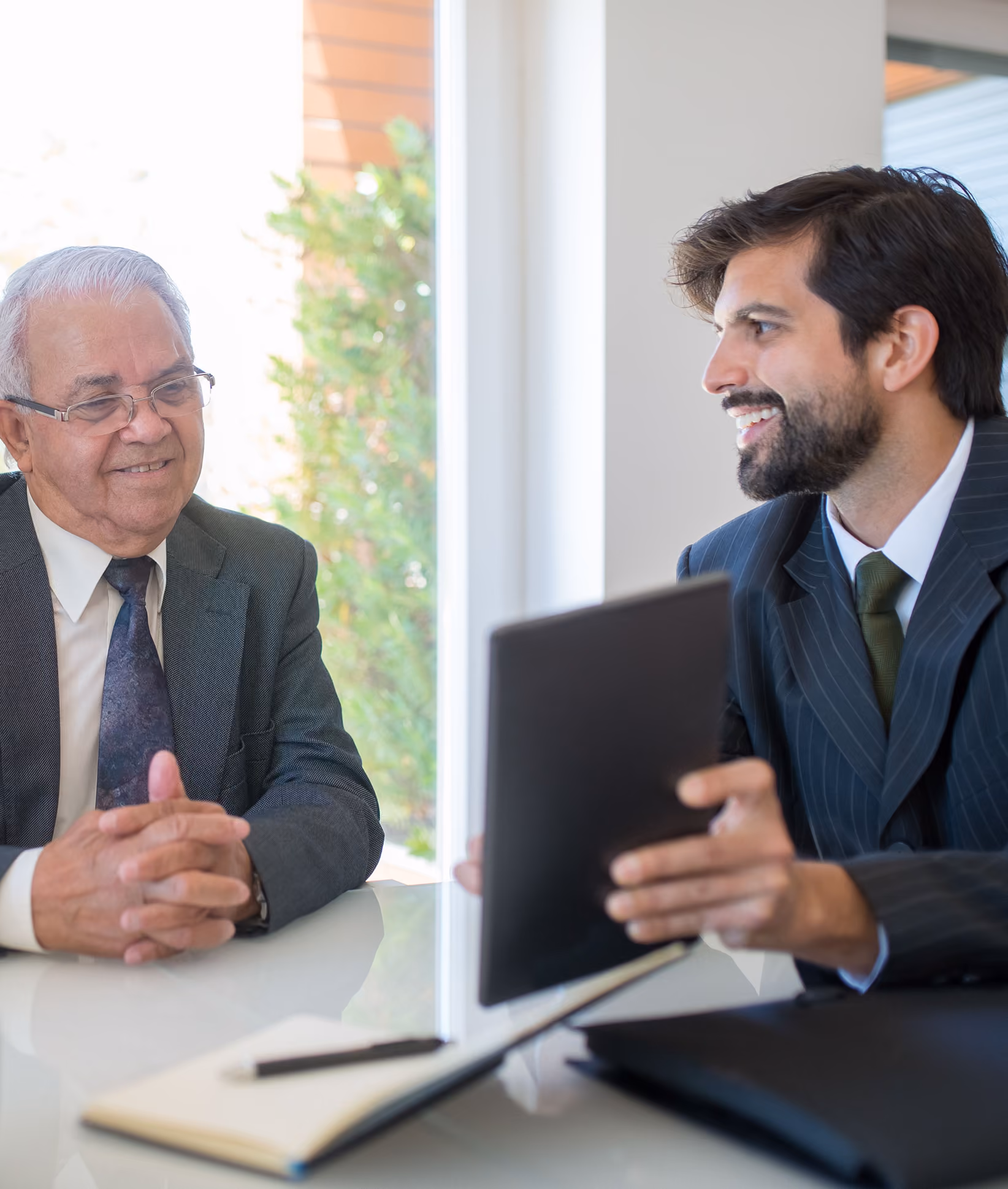 Two businessmen sitting at a table in an office, one older man listens while a younger man holds a tablet and smiles.