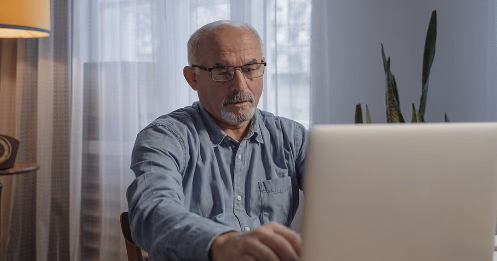 Older man with glasses and a gray beard seated indoors, using a laptop, with sheer curtains and a lit lamp visible in the background.
