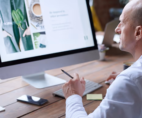Man sitting at a desk working on a desktop computer displaying a webpage, holding a stylus pen, with a smartphone and sticky notes on the desk.