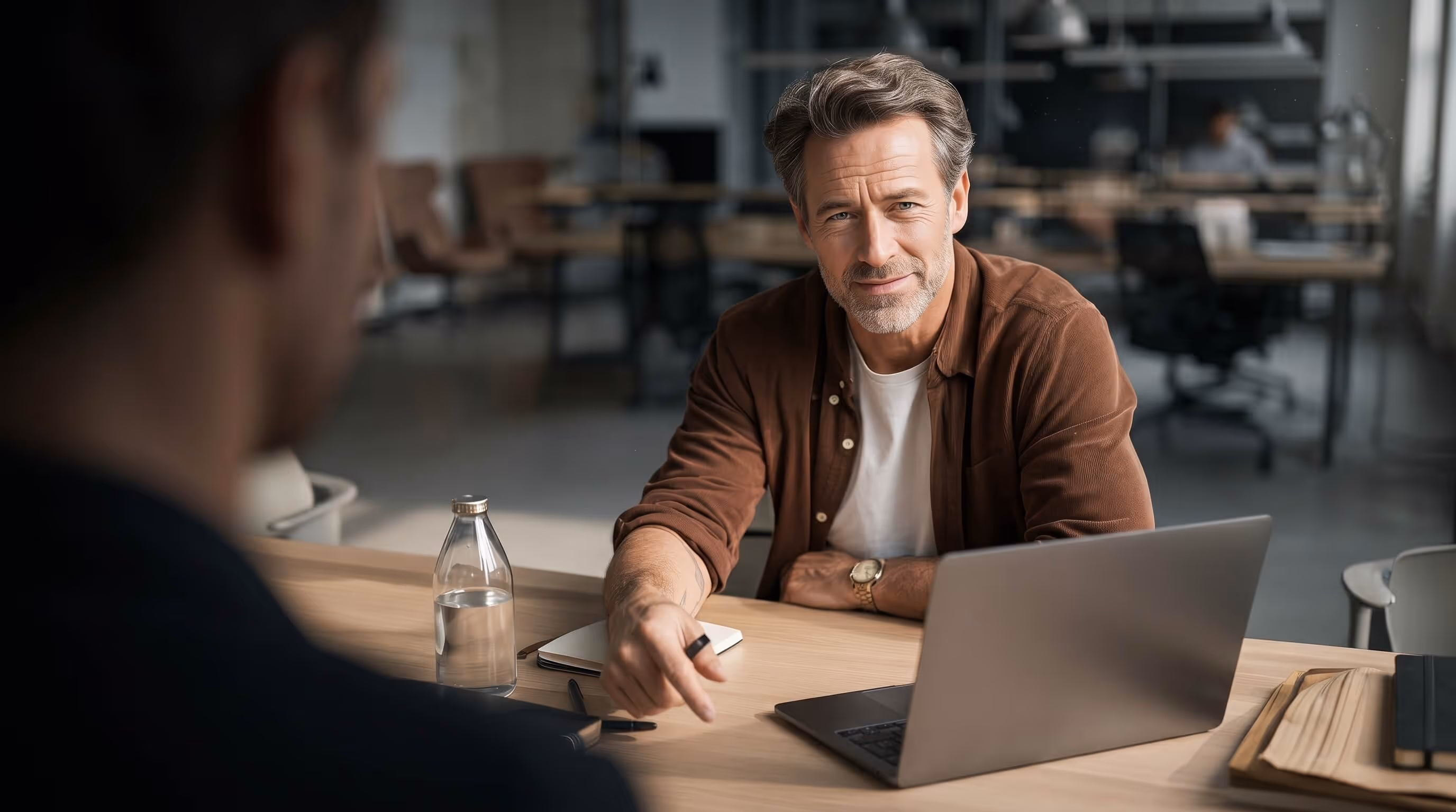 Man with gray hair and a beard pointing at a laptop during a conversation in an office setting.